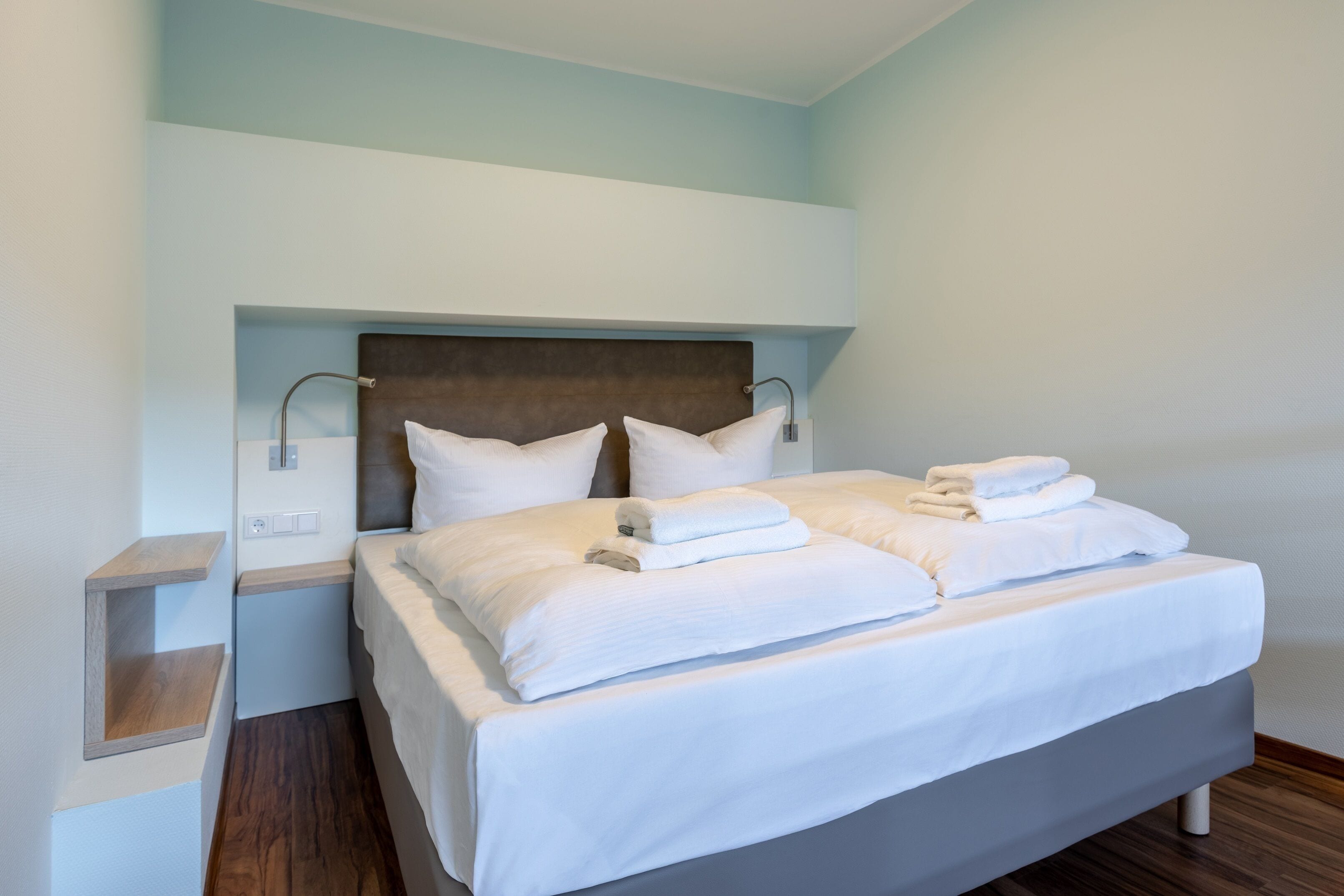 A neatly made bed with white linens and towels, a wooden headboard, and a bedside table with a lamp.