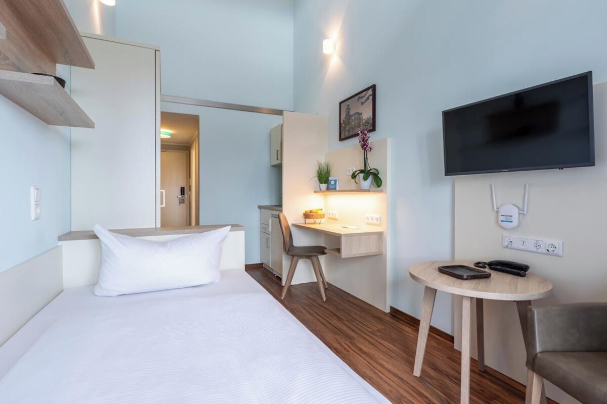 A hotel room with a bed, a desk, a chair, a TV, and a small table.