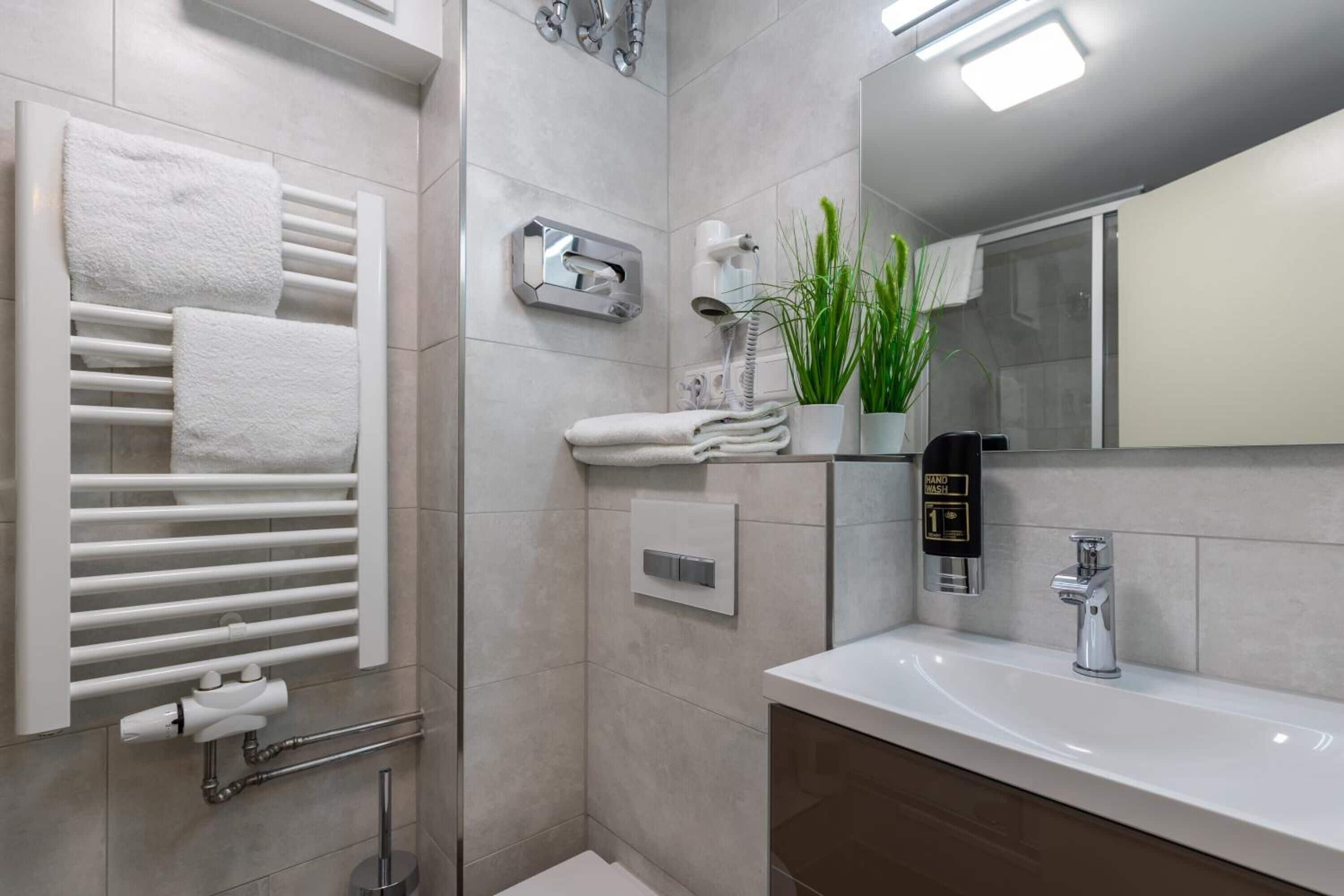 A modern bathroom with a white sink, a towel rack with folded towels, a handheld showerhead, and a potted plant.