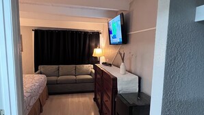 A compact living space with a sofa, a small table, a TV mounted on the wall, and a door leading to another room. - Regency Inn (Perry)