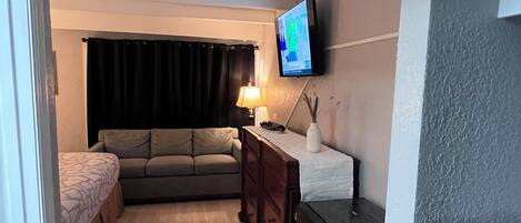 Un espacio habitable funcional con sofá, una mesita, televisor montado en la pared y una puerta que da a otra habitación.