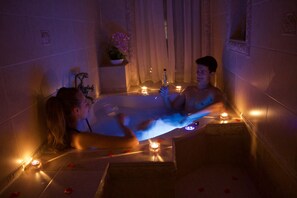 Un couple dans une baignoire, entouré de bougies et de pétales de rose, avec un mur carrelé et une plante en pot en arrière-plan.
