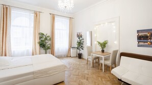 A hotel room with two beds, a dining table, a chair, a potted plant, a mirror with lights, and a cityscape painting. - Luxurious 5-bedroom apartment with wellness amenities (Prague)