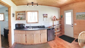 A compact kitchen with wooden cabinets, a sink, and a refrigerator. There is a window with a view of the landscape, and a door leading to another room. - Cozy Cabin in Kettle Valley (Rock Creek)