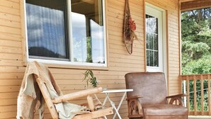 A wooden porch with a rocking chair and a leather chair, a small table, and a window with a view of greenery. - Cozy Cabin in Kettle Valley (Rock Creek)