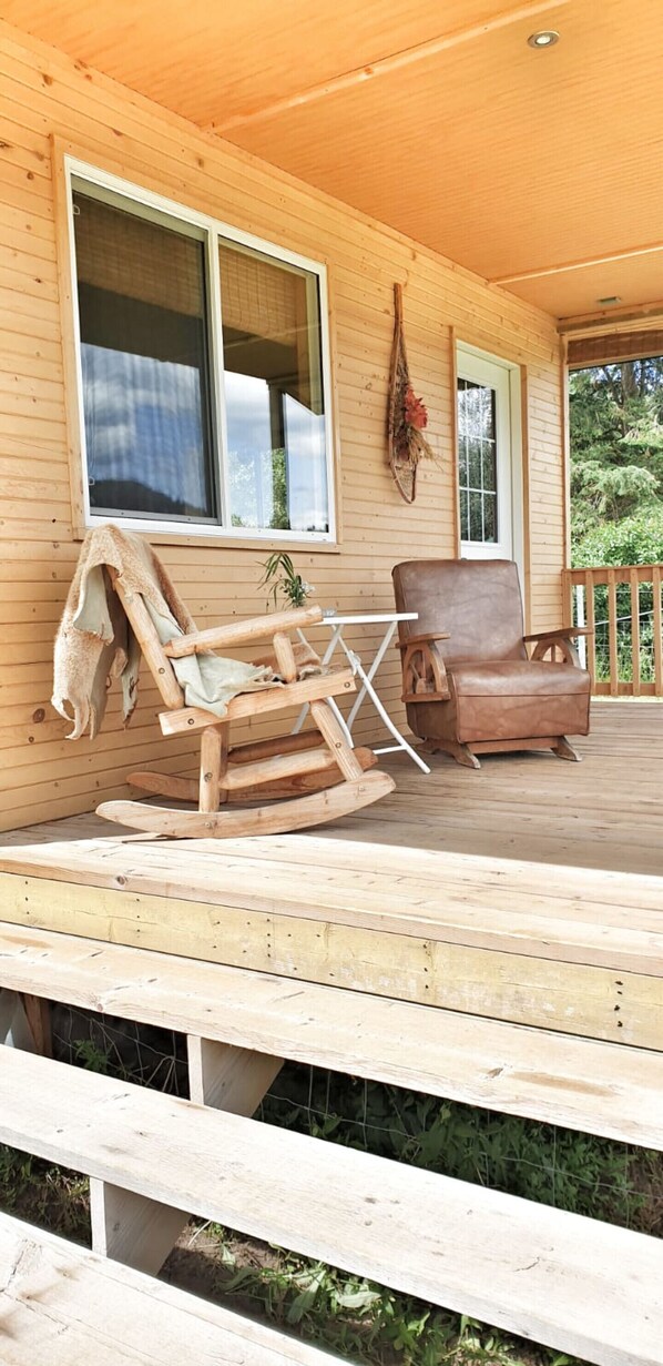 A wooden porch with a rocking chair and a leather chair, a small table, and a window with a view of greenery.