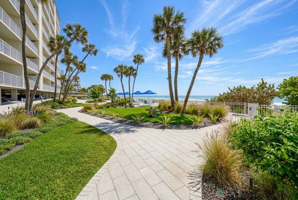 A paved walkway with palm trees, a pool, and a building with balconies. - Sand Castle 507: 2BD/2BA Beachfront Oasis (Indian Shores/Redington Shores)