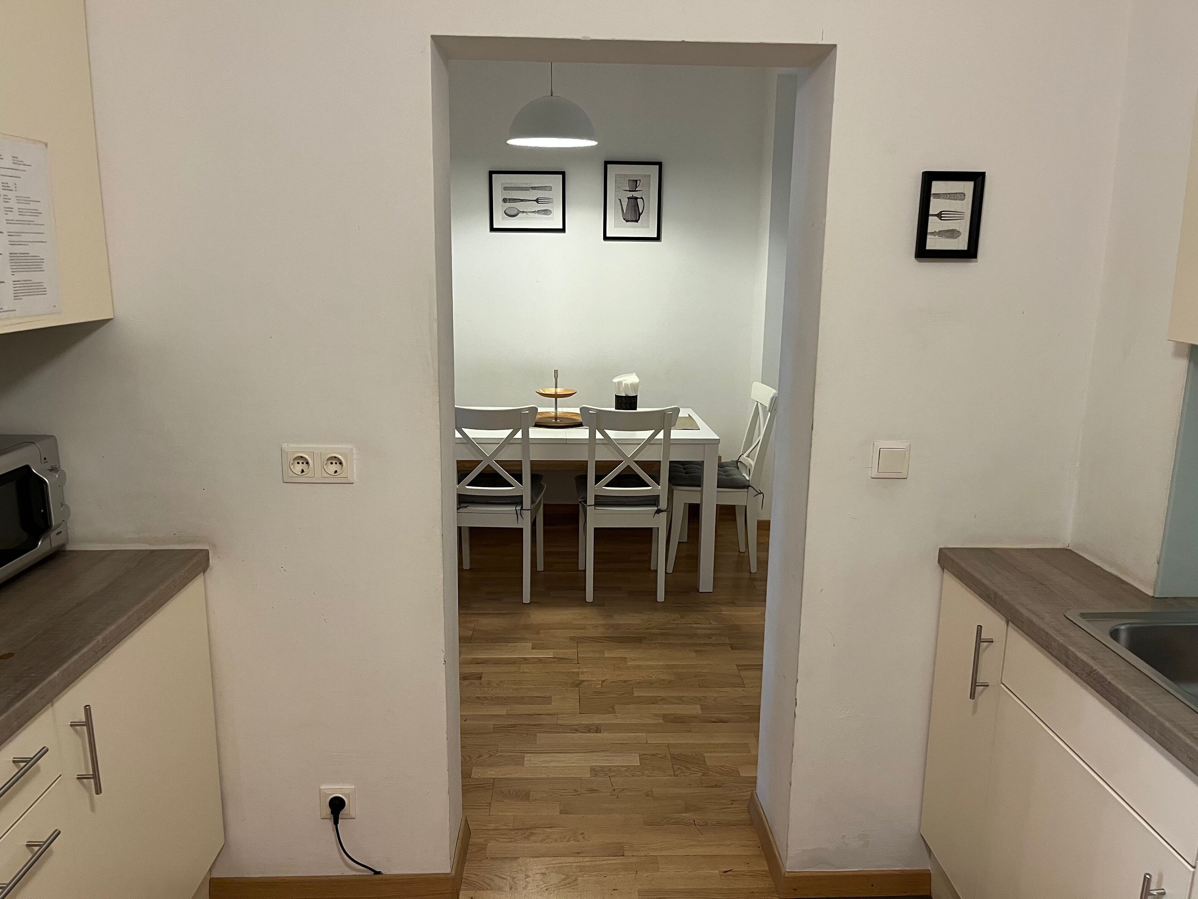 A narrow hallway leading to a dining area with a table and chairs, a hanging light fixture, and framed pictures on the walls.