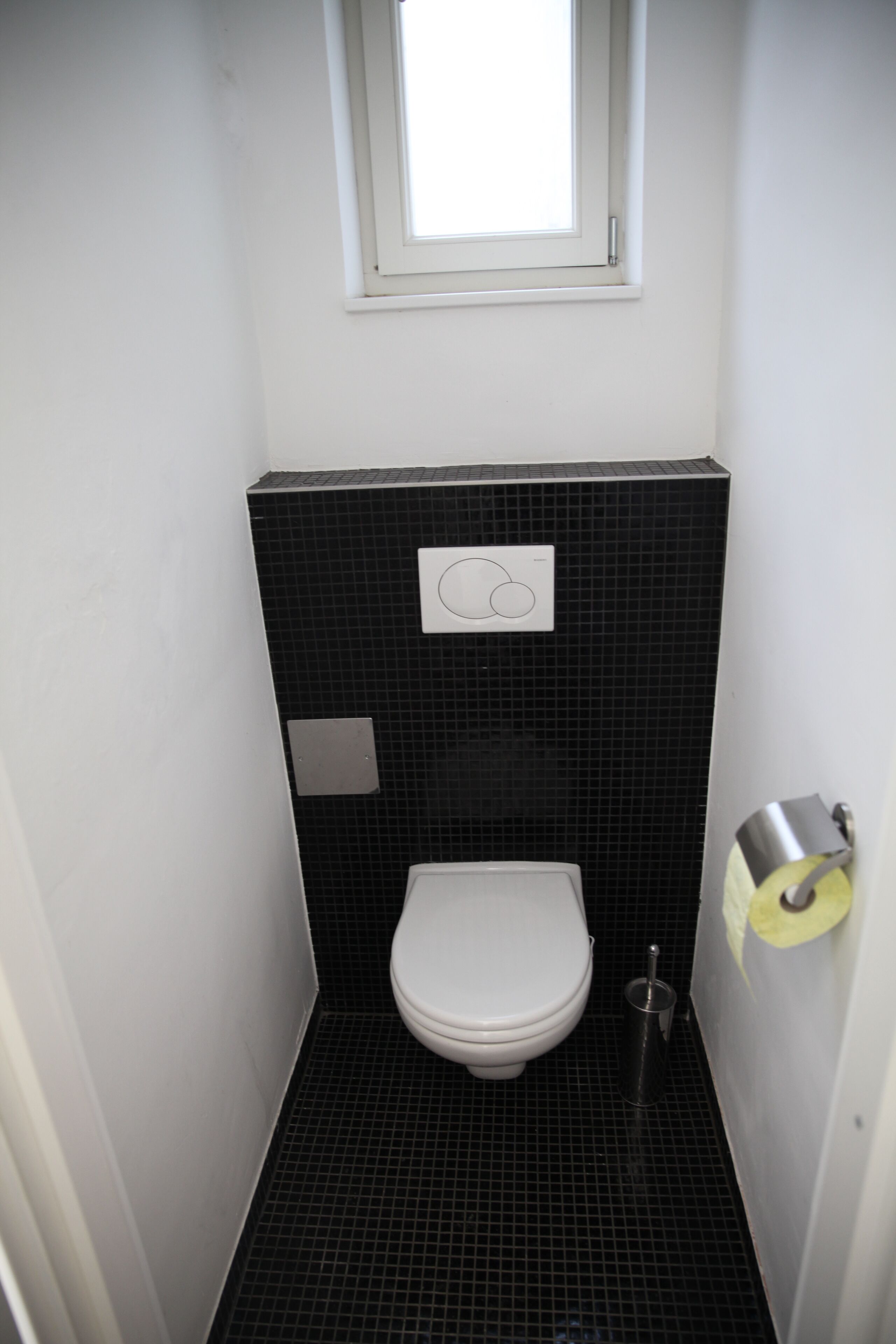 A small, narrow bathroom with a white toilet, a black tiled wall, and a window above the toilet.