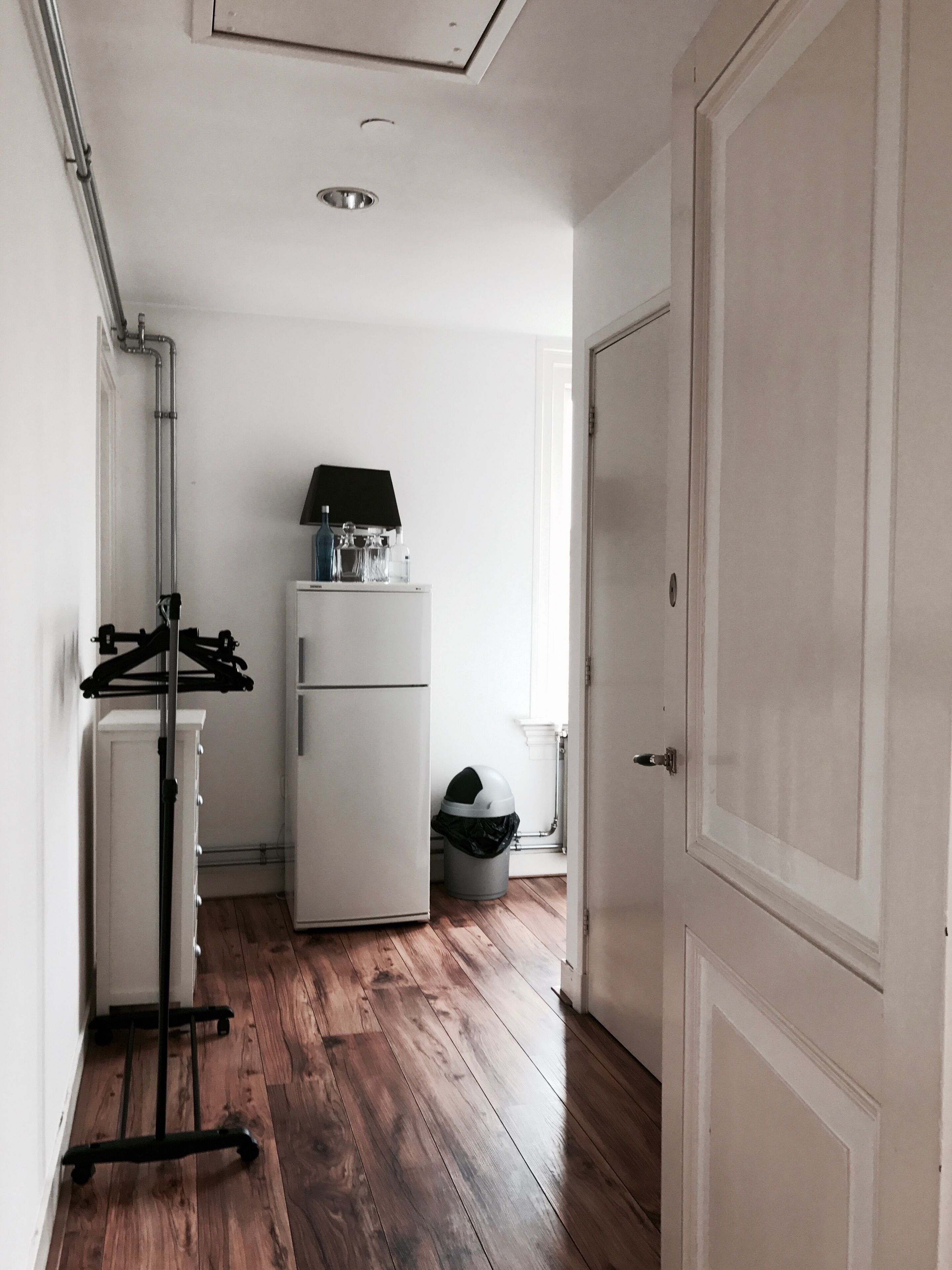 Un couloir étroit avec un réfrigérateur blanc, une petite poubelle et un porte-manteau.