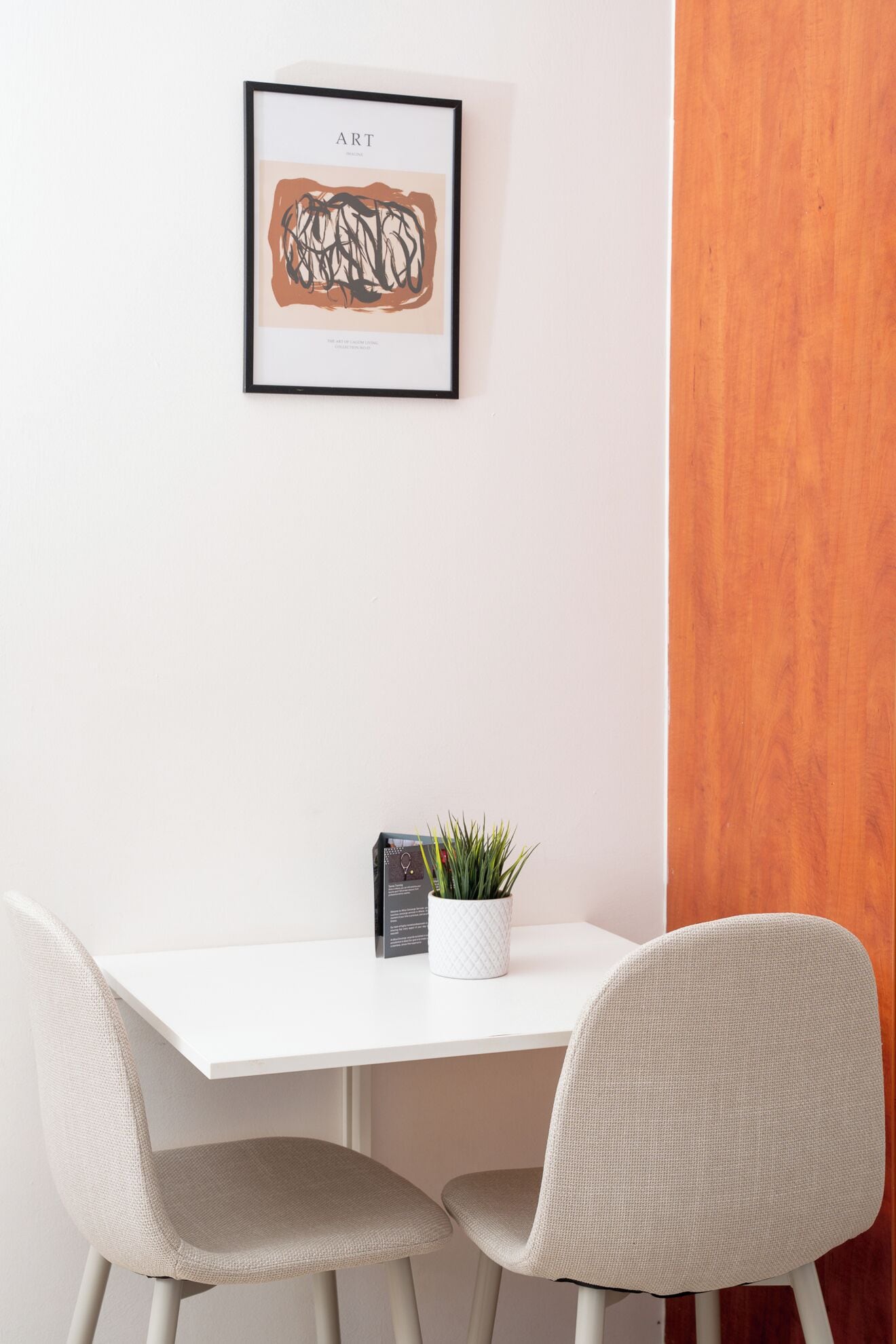 A white table with two chairs, a framed artwork above it, and a small potted plant on the table.