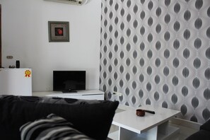 A modern living room with a white sofa, a flat-screen TV, a white coffee table, and a patterned wallpaper.