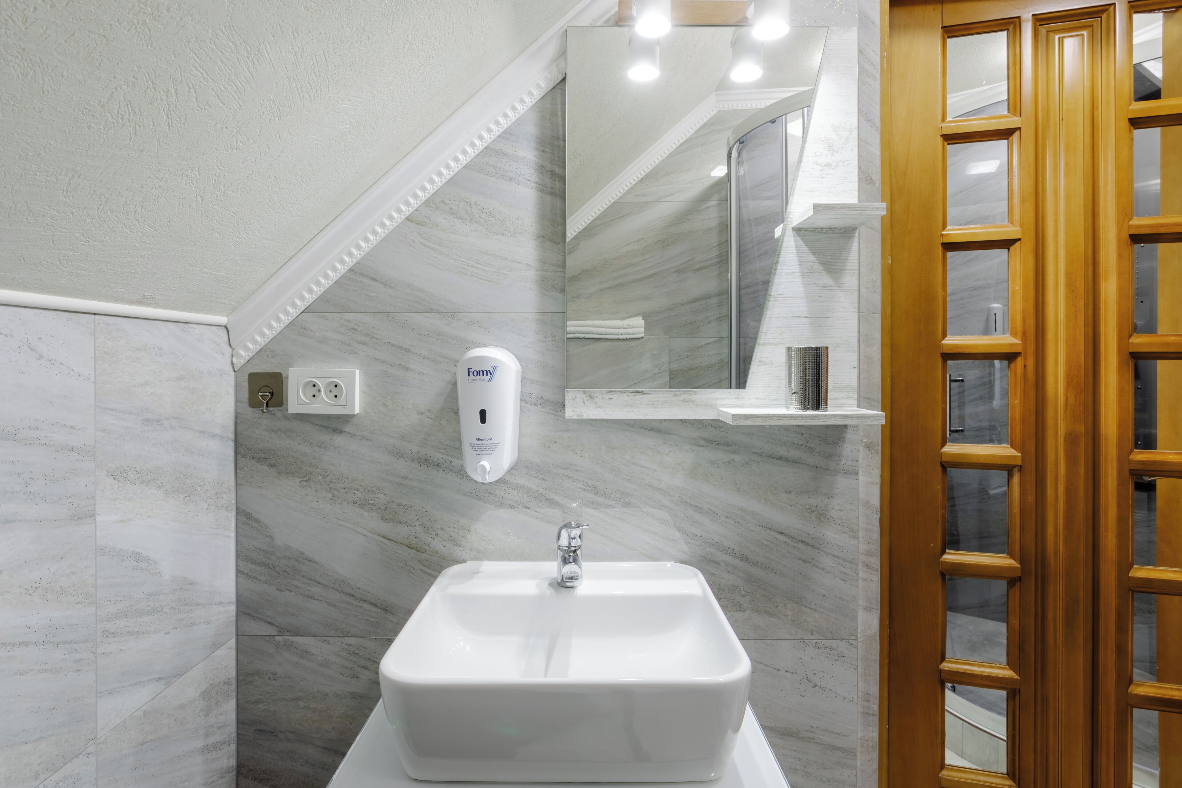 Un baño con un lavamanos blanco, paredes de mármol, un espejo y una puerta de madera.