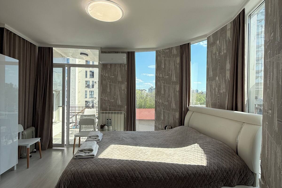 Een moderne slaapkamer met een groot bed, een bureau en een balkon met uitzicht op gebouwen.