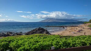 Uma praia com costa rochosa, céu azul claro e uma montanha ao fundo.
