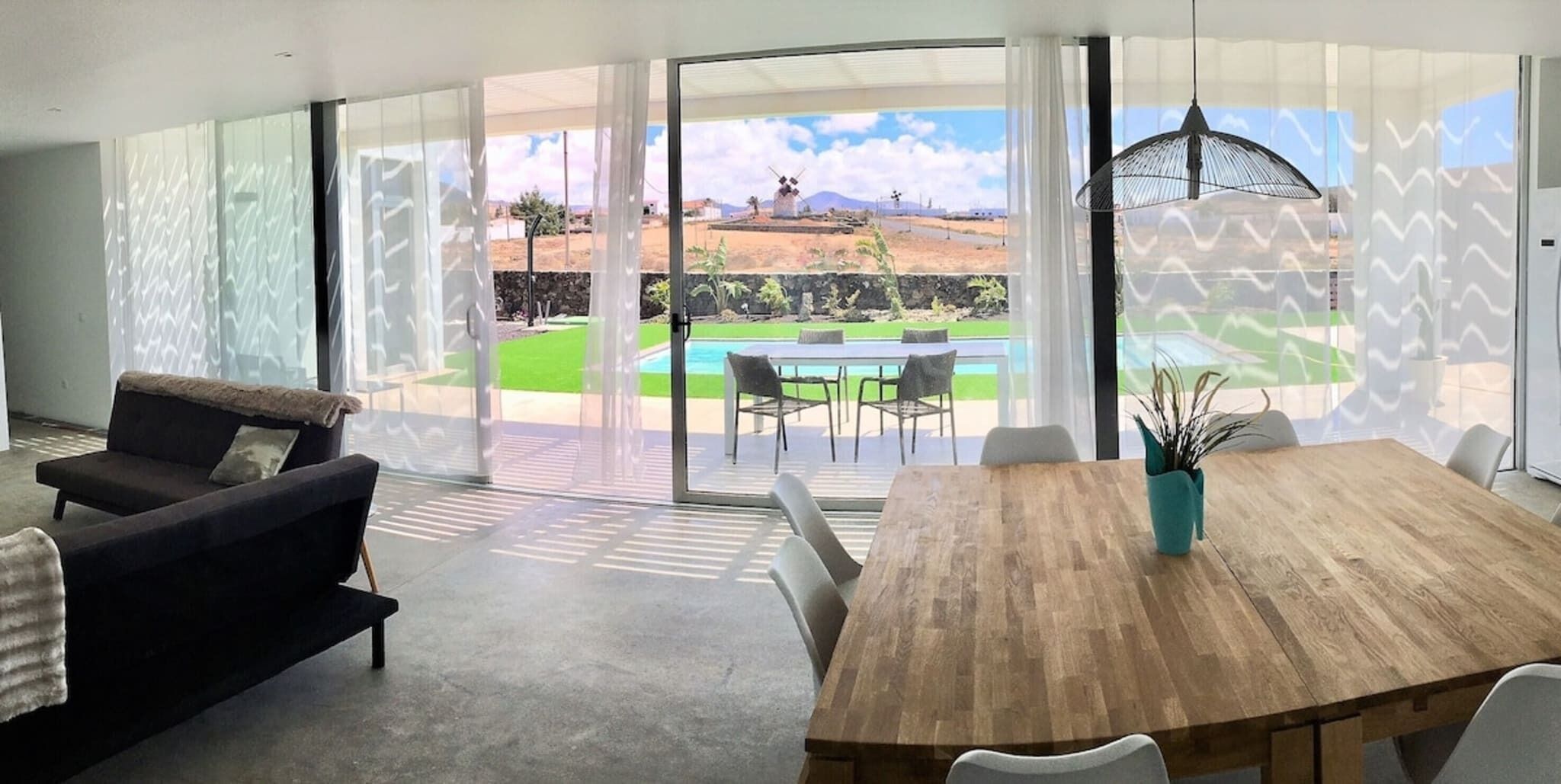A modern living room with a sofa, a wooden dining table, and a view of a swimming pool and windmills.