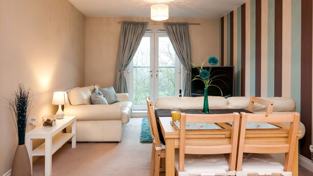 A modern living room with a sofa, a dining table with chairs, a lamp, and a view through a large window.