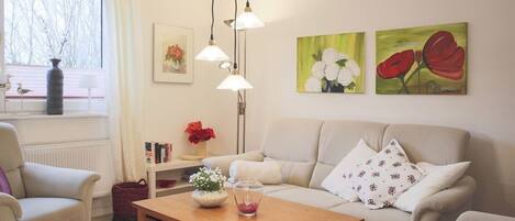 A cozy living room with a sofa, a wooden coffee table, and a bookshelf. There are paintings on the wall and a window with curtains.