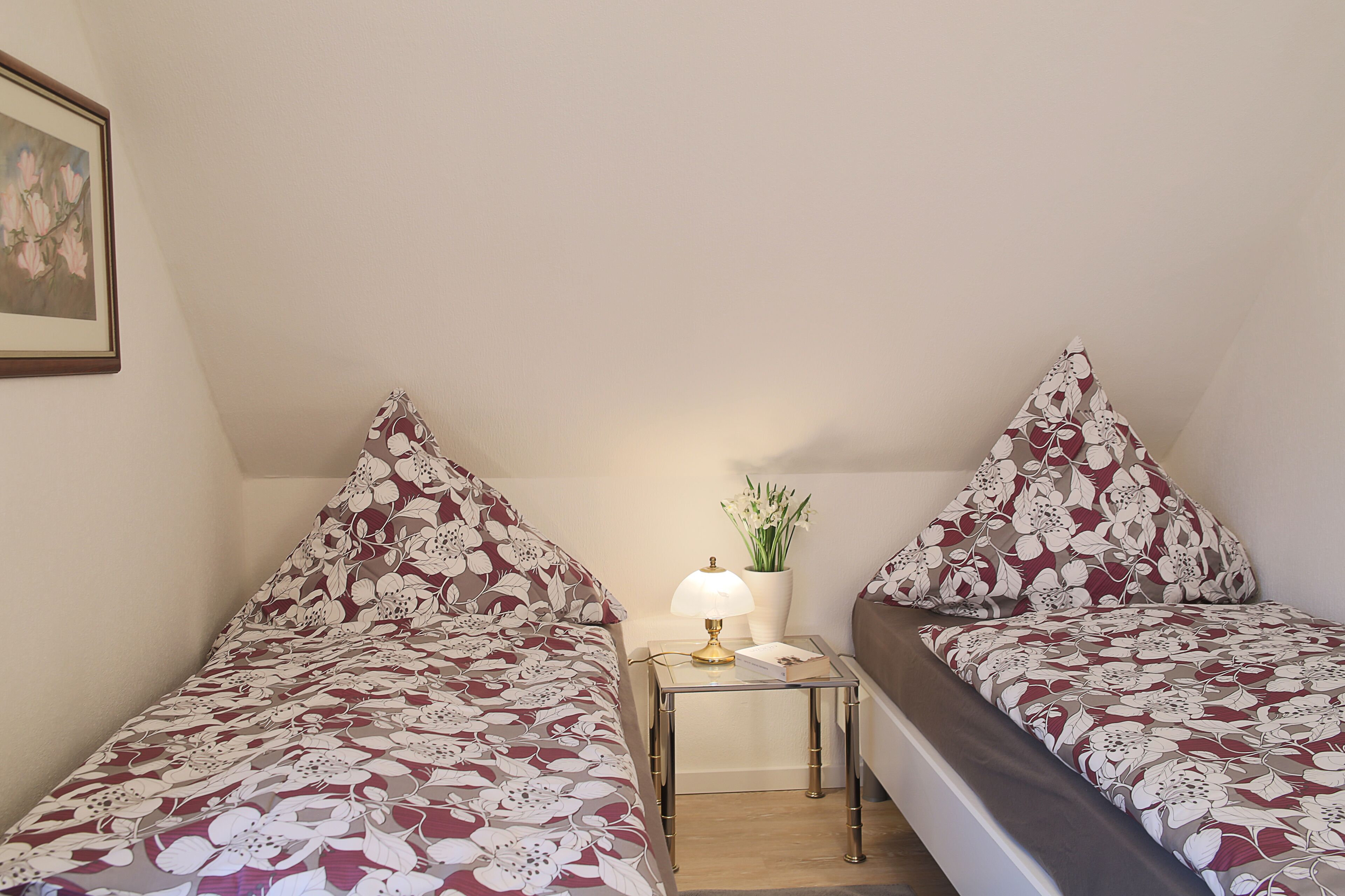 Une petite pièce triangulaire contenant deux lits, chacun recouvert d’une couvre-lit à motifs floraux. Une table de chevet avec une lampe et un vase de fleurs se trouve entre les deux lits.