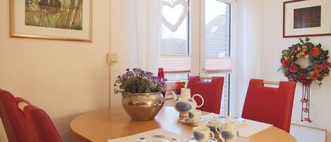 A dining area with a table set for two, a vase of flowers, and a heart-shaped decoration on the window.