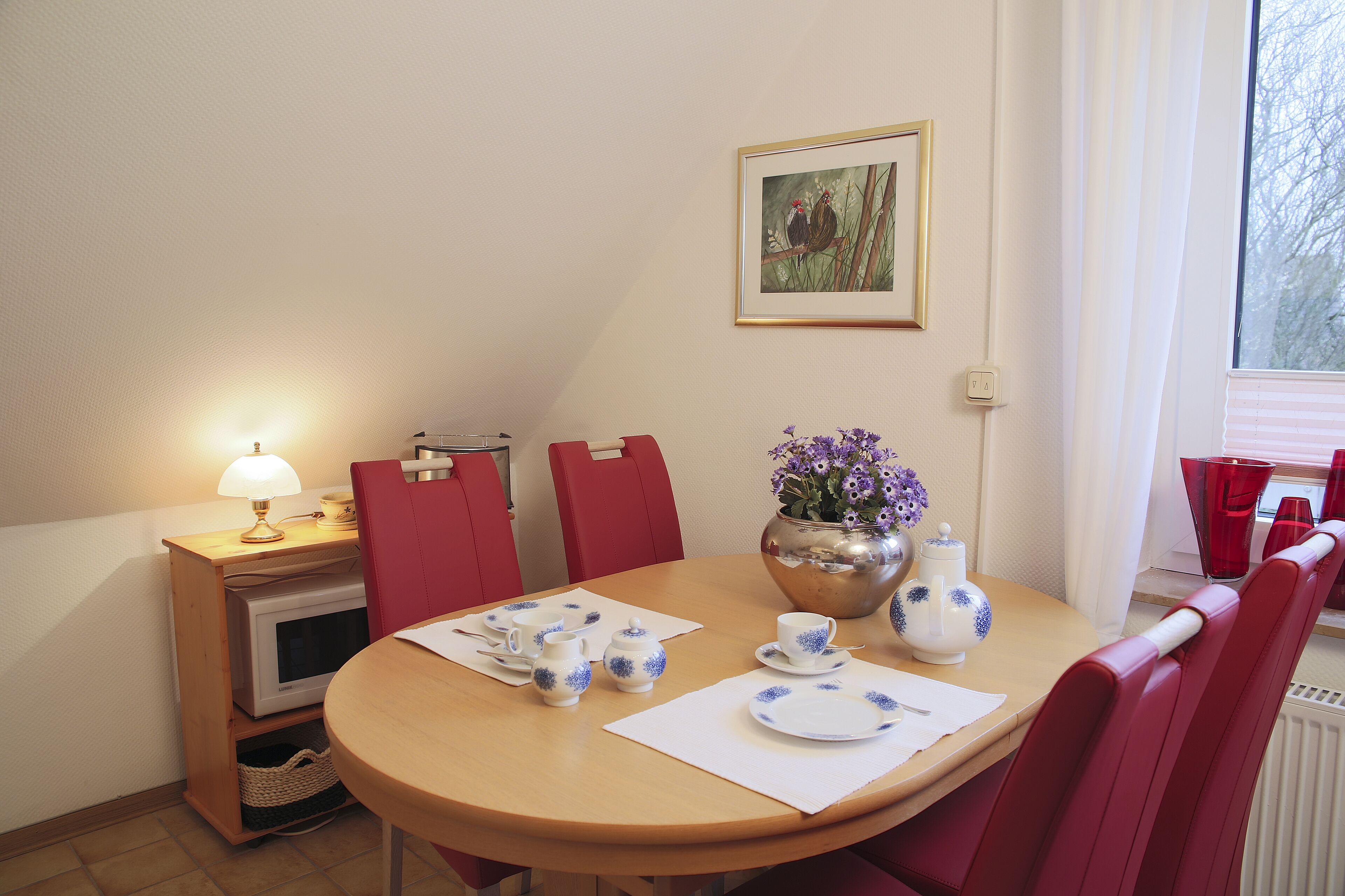 Un coin repas avec une table en bois dressée pour le thé, des chaises rouges, un petit coin cuisine et une œuvre d’art encadrée accrochée au mur.