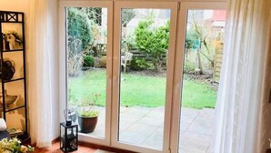 Una sala de estar con alfombra roja, una mesa de centro de vidrio y una estantería. Hay una puerta corrediza de vidrio que da a un jardín con césped y plantas en macetas.