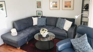 Una sala moderna con un sofá seccional gris oscuro, una mesa de centro de vidrio y un sillón azul. Hay obras de arte enmarcadas en la pared y una lámpara blanca sobre una mesita auxiliar.