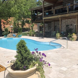 Un área de piscina con muelle de madera, plantas en macetas y un edificio de dos pisos con balcón.