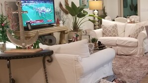 Een woonkamer met een televisie, witte banken, een salontafel, sierplanten en een lamp.