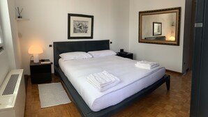 Una cama bien hecha con ropa de cama y toallas blancas, una lámpara de noche, un cuadro en la pared y un espejo grande.