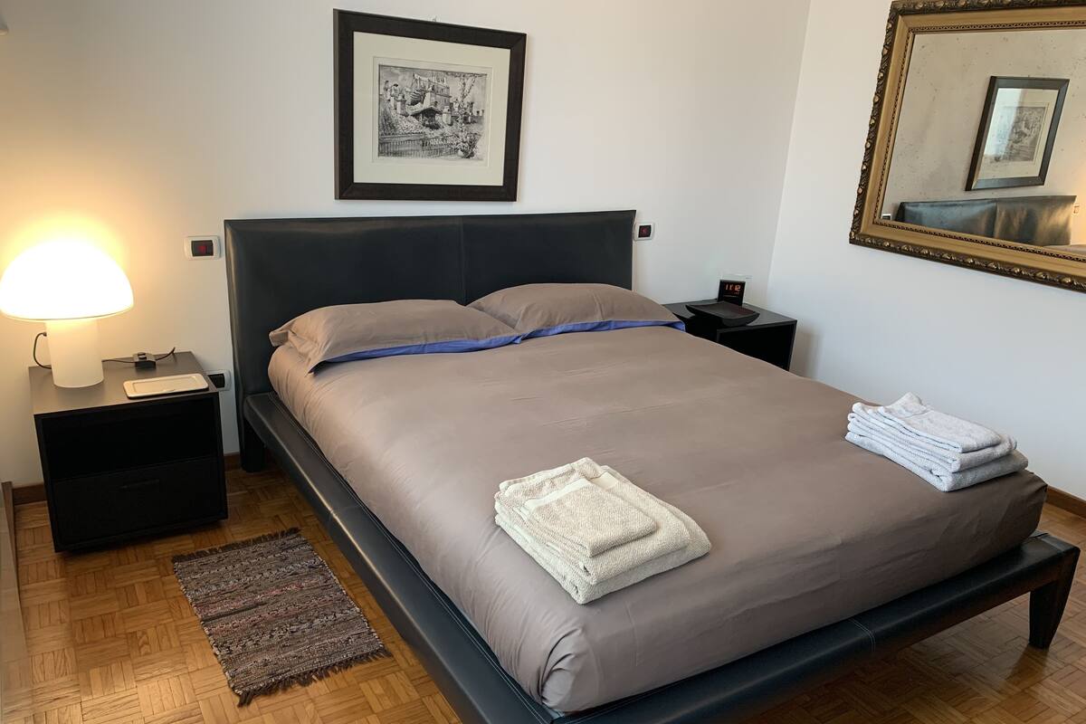 Una cama bien hecha con una colcha gris, dos almohadas y toallas dobladas a un lado. Hay una mesita de noche con una lámpara y un reloj pequeño. Un cuadro enmarcado cuelga en la pared sobre la cama.