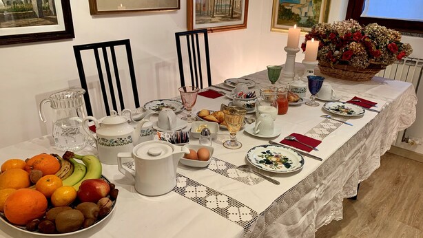 Una mesa de comedor dispuesta con frutas, huevos y diferentes vajillas, rodeada de sillas y con cuadros en la pared.