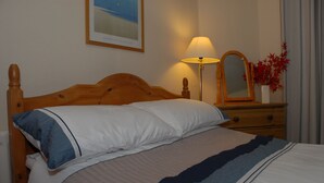A neatly made bed with a wooden headboard, a bedside lamp, a mirror, and a vase with red flowers on a dresser. - Keswick, Garden Apartment for 2. Excellent location.Convenient for all amenities (Keswick)