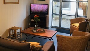 Ein Wohnzimmer mit einer Couch, zwei Korbstühlen, einem Couchtisch mit Blumenvase, einem Fernseher und einem großen Fenster mit Blick ins Freie.