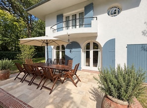 A two-story house with a patio area featuring a wooden table and chairs, a large umbrella, and potted plants.