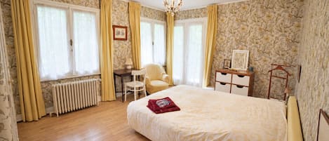 A hotel room with a large bed, a desk, a chair, a radiator, and a chandelier.