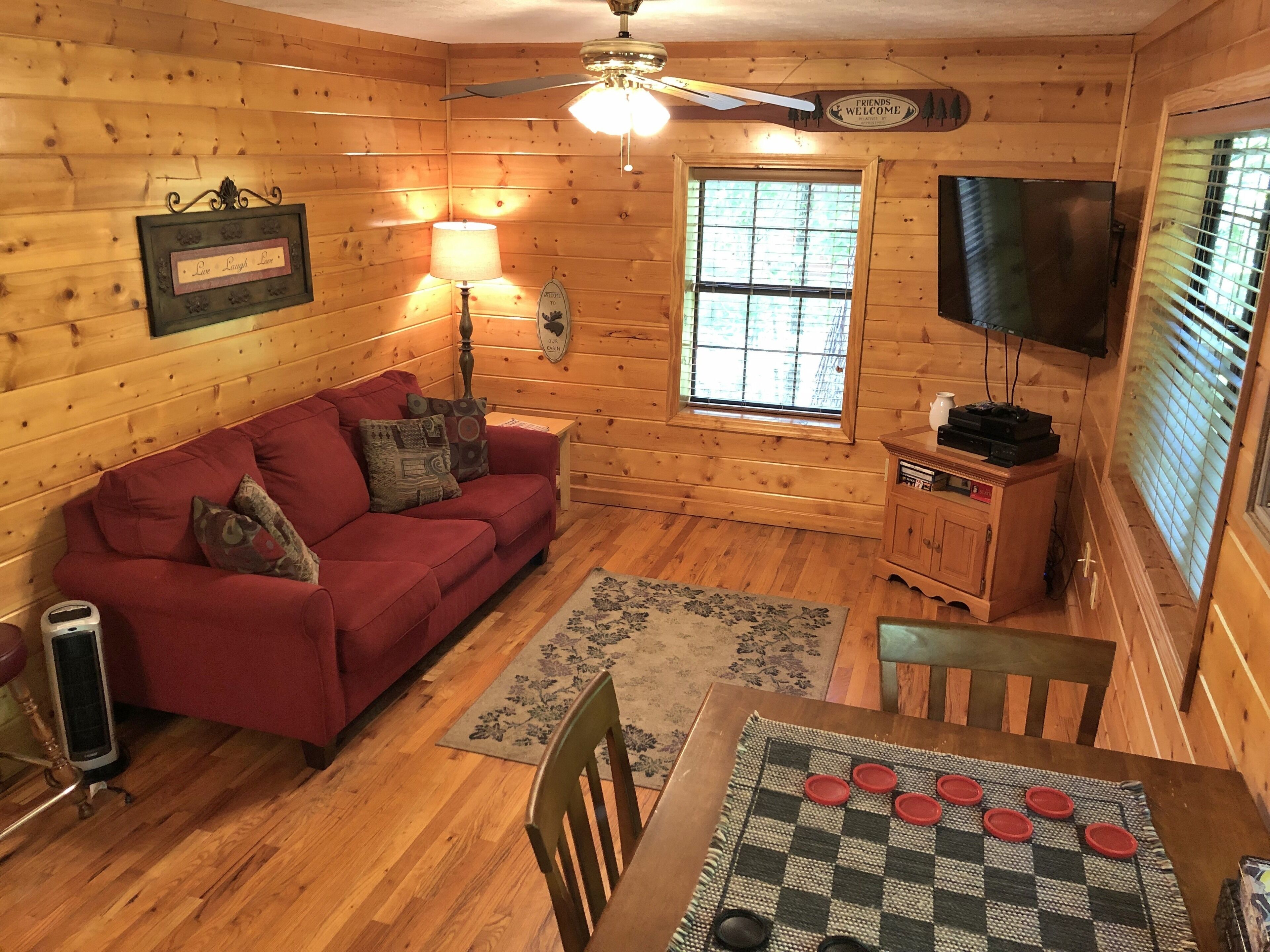 A cozy cabin interior with a red sofa, wooden walls, a ceiling fan, a television, and a table with a checkered table runner.