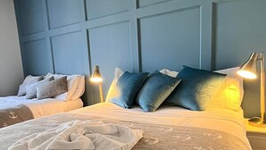 Habitación de hotel con una cama grande, una cama más pequeña y un cabecero con diseño de cuadrícula. Hay dos lámparas de noche y una manta con motivos de anclas sobre la cama.