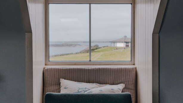 Una habitación con un ventanal grande, un banco con cojines y vistas a un espacio verde y un edificio al otro lado del agua.