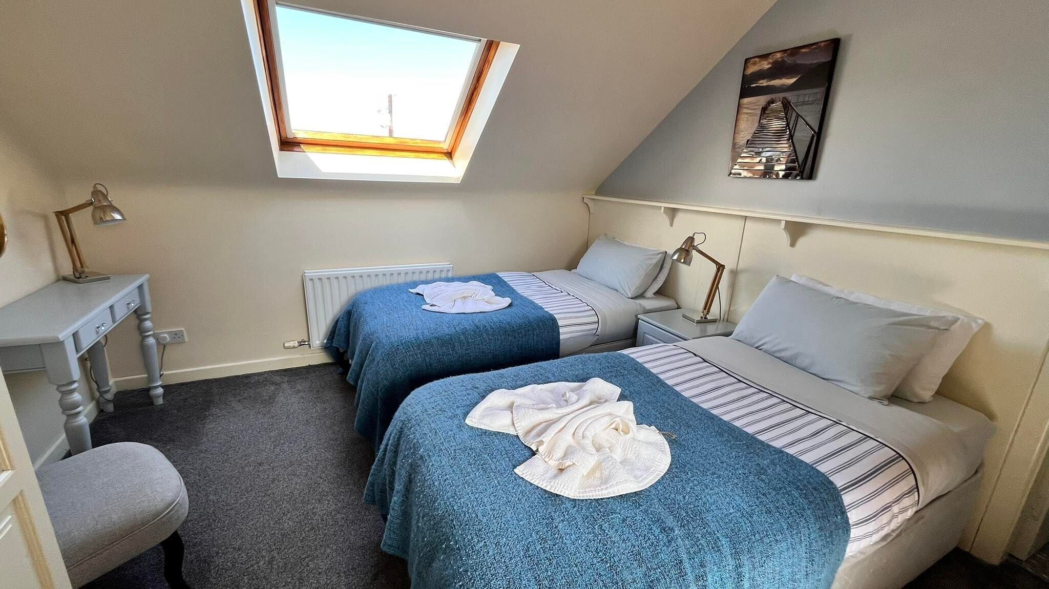 A room with two beds, a desk, a chair, a radiator, and a skylight.