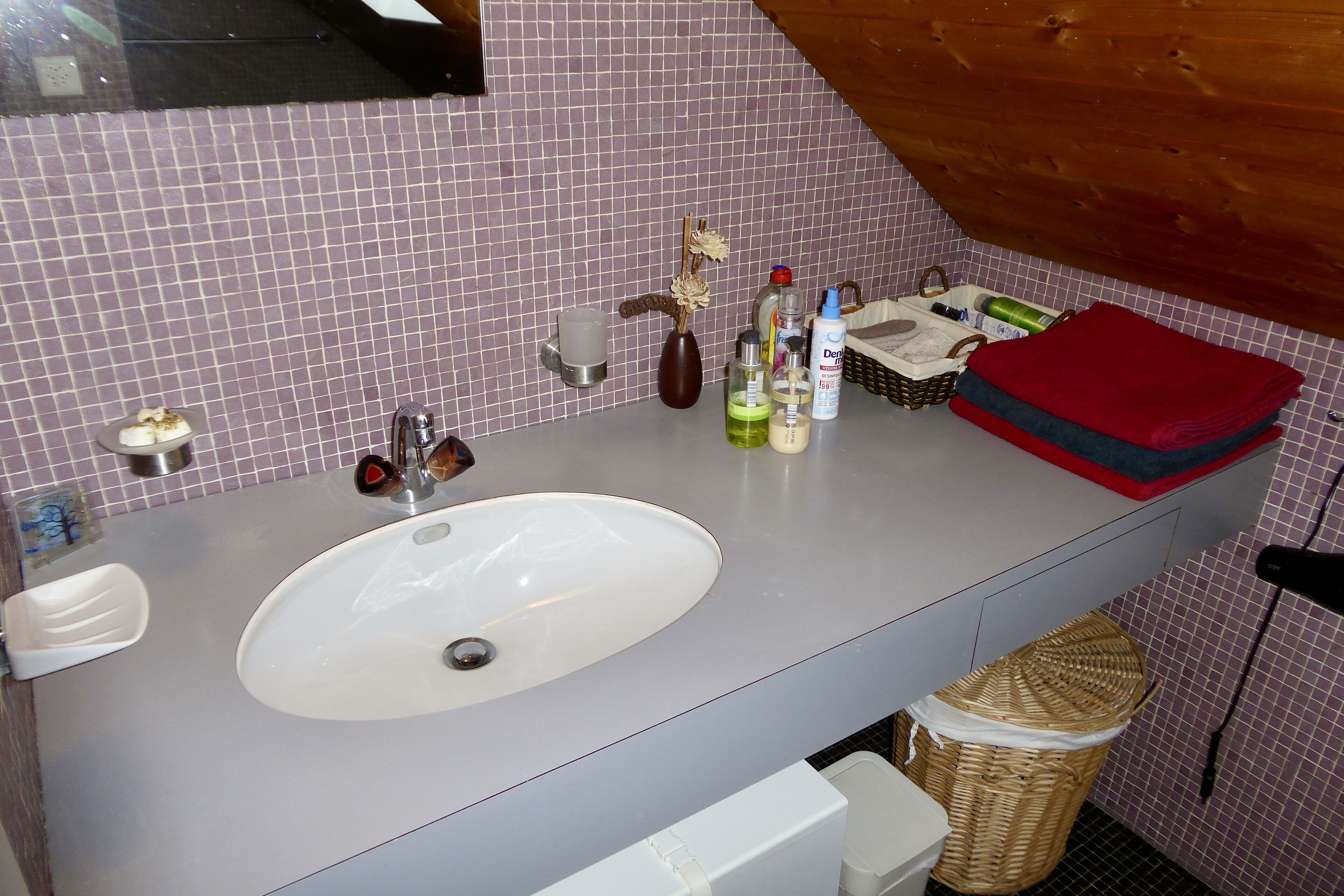 Une salle de bain avec un lavabo blanc, un miroir et un mur recouvert de carreaux. On y trouve des articles de toilette sur le comptoir et un panier.