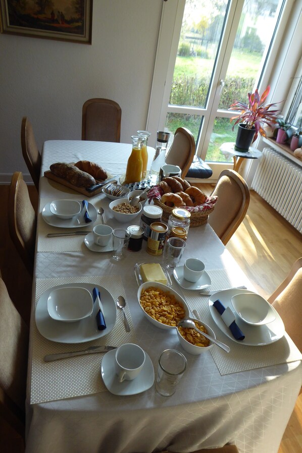 Mesa de desayuno preparada con platos, tazones, tazas y diversos alimentos para el desayuno.