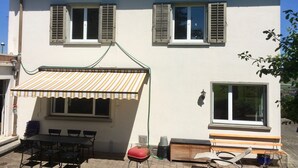 Ein zweistöckiges Haus mit Ziegeldach, einer überdachten Terrasse mit Streifenmarkise und einem Grill.