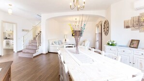 Un comedor amplio con una mesa larga, sillas blancas y una lámpara de araña. Una escalera conduce a otra habitación, y se ve una cocina al fondo.