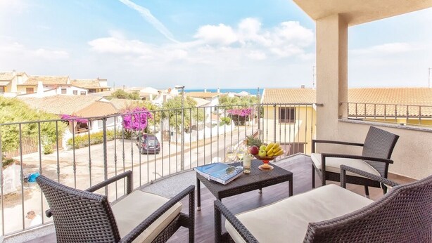 Un balcón con mobiliario de mimbre, una mesa con frutero, y vista a una zona residencial y el mar.