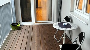Una terraza de madera con una pequeña mesa redonda y dos sillas, una puerta de vidrio corredera y una ventana con cortinas.