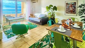 A modern living room with a dining area, large windows overlooking the sea, a sofa, a dining table set with plates and a bottle, and a green chair.