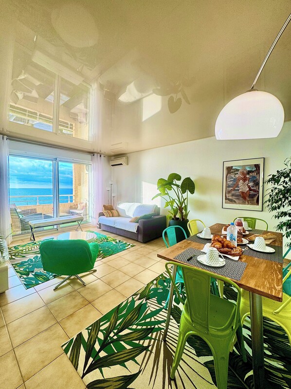 A modern living room with a dining area, large windows overlooking the sea, a sofa, a dining table set with plates and a bottle, and a green chair.