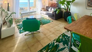 A spacious living area with a large window overlooking the sea, a cozy seating area with a sofa and chairs, and a dining table with chairs.