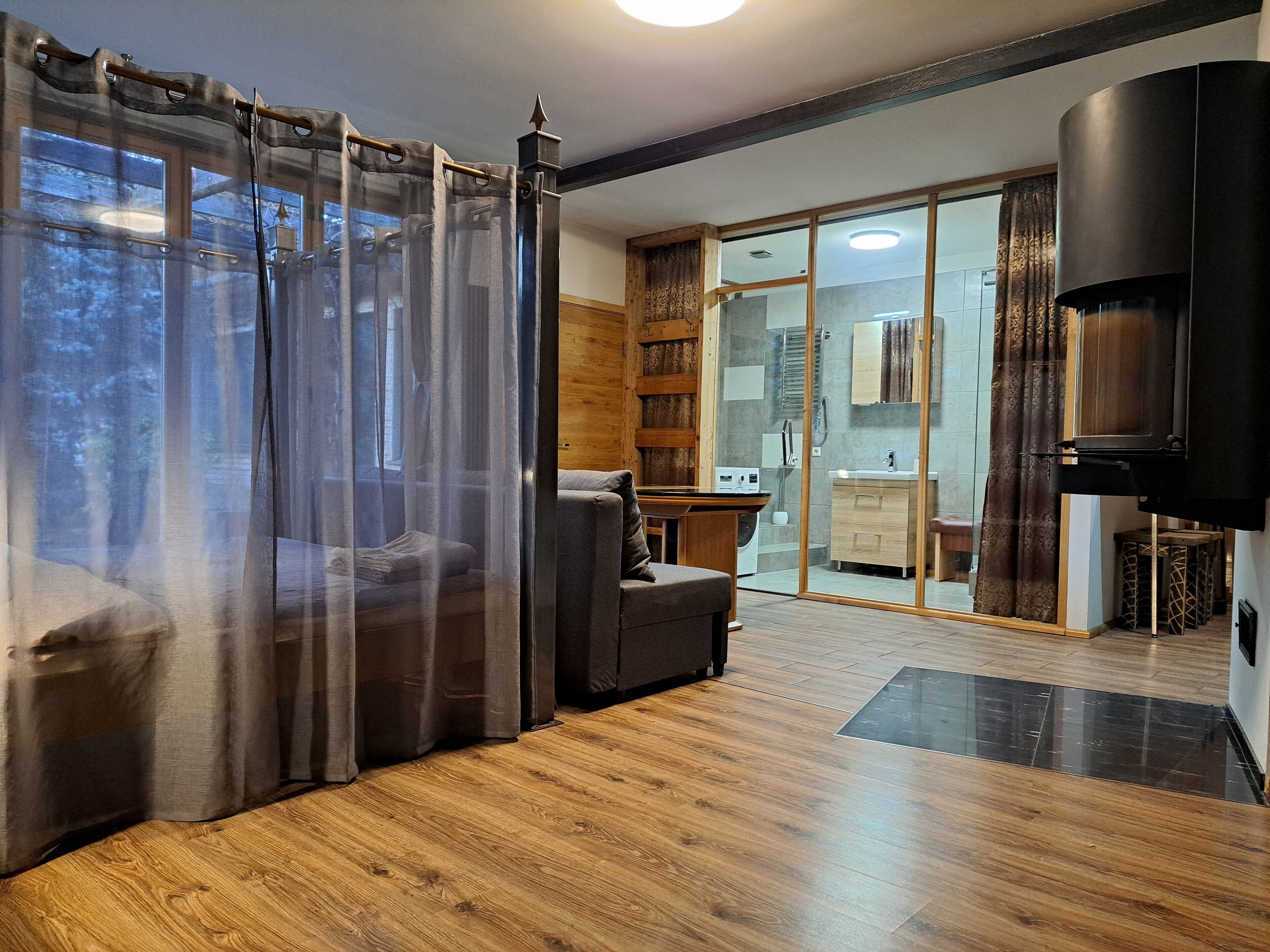 A modern living room with a wooden floor, a grey sofa, a TV, and a sliding glass door leading to a bathroom.